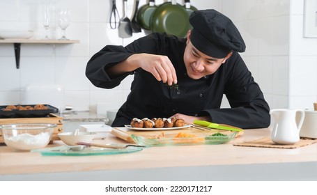 Asian Young Handsome Chef Man, In Black Uniform, Smiling While Garnishing Seaweed On Japanese Food Called Takoyaki In Plate Ready To Serve From Hot Pan With Ingredients On Table At Kitchen Restaurant