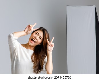 Asian Young Happy Cheerful Long Hair Female Model In Casual Outfit Standing Smiling Holding Hands Up Showing Two Fingers Peace Sign At Head In Studio Photography Set In Front Lighting Soft Box Lamp.