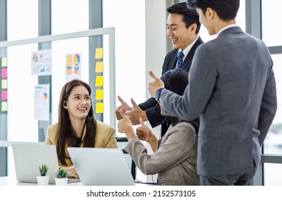 Asian young happy customer service operator helpdesk employee staff with microphone headset sitting smiling at working desk while male female colleagues showing thumb up admire complimenting good job.