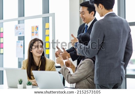 Asian young happy customer service operator helpdesk employee staff with microphone headset sitting smiling at working desk while male female colleagues showing thumb up admire complimenting good job.