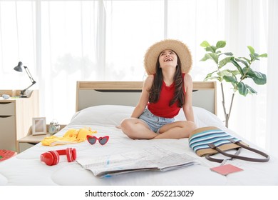 Asian Young Happy Female Traveler Wears Big Hat Laughing Smiling Sitting Relaxing On Bed Preparing Personal Stuff For Sea And Beach Trip With Handbag Notebook Sunglasses Bikini Paper Map And Headset.