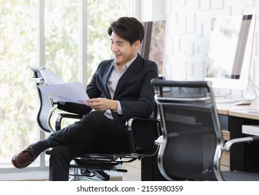 Asian Young Happy Handsome Confident Successful Professional Male  Businessman Entrepreneur Wears Black Formal Suit Sitting Smiling On Chair In Office Reading Analyzing Report Chart Document In Hands.