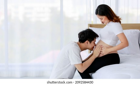 Asian Young Happy Loving Supporting Comforting Soothing Husband Sitting Smiling Kissing Lovely Unborn Baby In Beautiful Pregnancy Wife Naked Big Belly Tummy Stomach Sitting On Bed In Bedroom At Home.