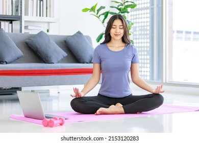 Asian young happy peaceful calm female model in casual sporty outfit sitting crossed legs in lotus position on yoga mat learning studying online meditation class via laptop computer in living room. - Shutterstock ID 2153120283