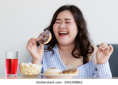 Asian young happy pretty friendly fat plus size woman sit smiling relaxing on gray fabric sofa while eating delicious chocolate donut in hand.