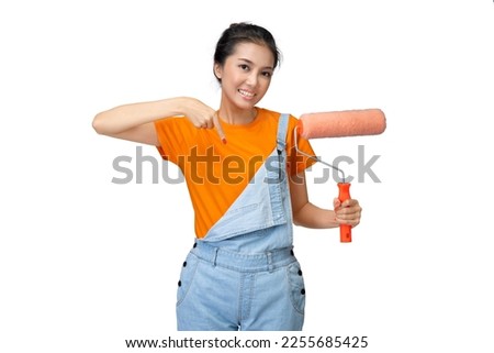 Asian young housewife holding a roller paint brush and pointing something isolated over white background