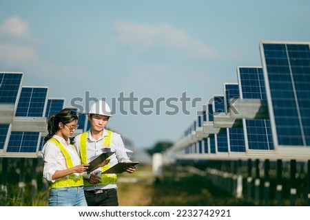 Asian Young Inspector Engineer male and female wearing protection helmet holding paperwork and board in hands walking between row of solar panel while checking operation in solar station