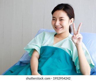 Asian Young Long Hair Happy Female Patient Wears Green Hospital Uniform Covered By Blanket Sit Lean On Blue Pillow On Bed Hold Hand Shows Victory Sign When Recovery And Good Condition In Private Room.