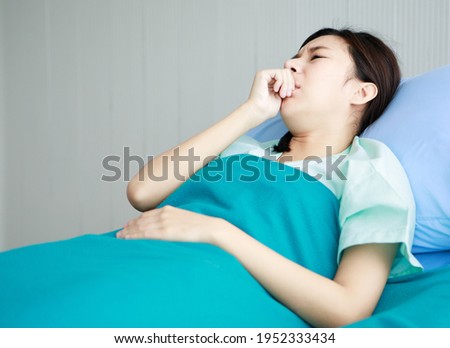 Asian young long hair sick ill girl patient wears green hospital uniform lay down on bed covered by blanket hold hand on chest sudden suffering from cough and pain in private ward room.
