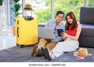 Asian Young Lovely Boyfriend And Girlfriend Traveler Couple Sitting Smiling On Carpet In Living Room With Trolley Luggage While Using Laptop Computer And Tablet Booking Vacation Summer Trip Online.