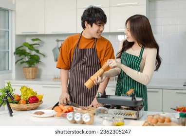 Asian young lover couple husband and wife in casual outfit with apron standing smiling man using hands massaging fresh meat while woman put pepper on chopping board in full decorated modern kitchen. - Powered by Shutterstock