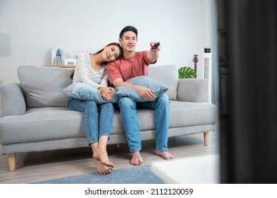 Asian young loving couple watch movie together in living room at home. Attractive romantic new marriage man and woman sitting on sofa, looking at TV show and having fun laughing together in house.