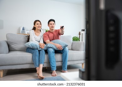 Asian Young Loving Couple Watch Movie Together In Living Room At Home. Attractive Romantic New Marriage Man And Woman Sitting On Sofa, Looking At TV Show And Having Fun Laughing Together In House.