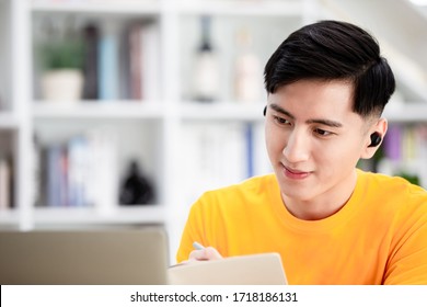 asian young male student use wireless earbuds to have online language courses and writes notes via laptop computer at home   