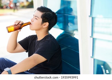 Asian Young Man Drinking Beer From Bottle While Sitting Outdoors