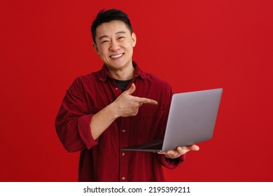 Asian Young Man Laughing And Pointing Finger At His Laptop Isolated Over Red Background