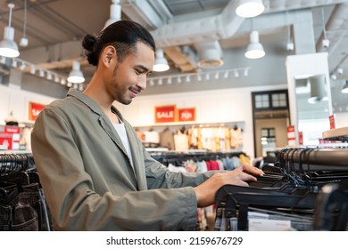 Asian Young Man Look And Choose Products Of Clothes In Shopping Mall. Attractive Happy Male Enjoy Walking In Department Store To Choose New T-shirt And Pants From Clothing Racks In Marketplace Center.