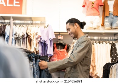 Asian Young Man Look And Choose Products Of Clothes In Shopping Mall. Attractive Happy Male Enjoy Walking In Department Store To Choose New T-shirt And Pants From Clothing Racks In Marketplace Center.