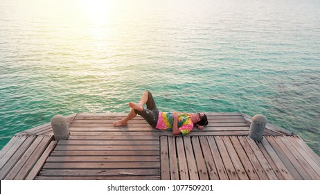 Asian Young Man Lying Down Under The Sun On Wood Bridge Near Blue Sea.