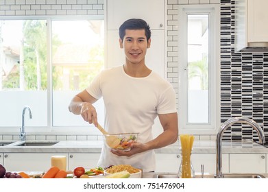 Asian Young Man Mixing Salad And Smiling While Cooking In Kitchen At Home. 