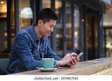 Asian Young Man At Outdoor Cafe, Using Mobile Phone, With Happy Smilie