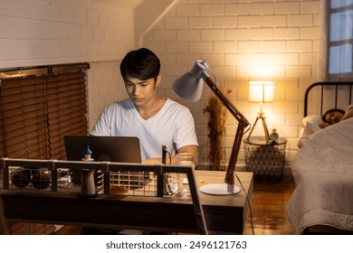 Asian young man overworking while his wife sleeping in bedroom at night. Attractive handsome businessman husband typing laptop computer working communicate online on table during bedtime in house. - Powered by Shutterstock