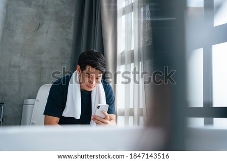 Asian young man relax in toilet and using smartphone.