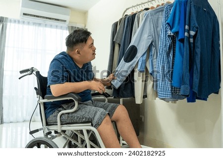 Asian young man sit on wheelchair and choosing clothes on closet rack. Attractive male patient person feeling happy and relax, then getting dressed in bedroom for morning lifestyles at house.
