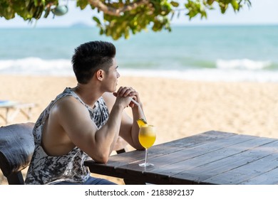 Asian young man sitting alone and drinking cocktail juice on the beach. Attractive handsome male sit on dinner table and looking at view at seaside enjoy holiday vacation trip in tropical sea island.