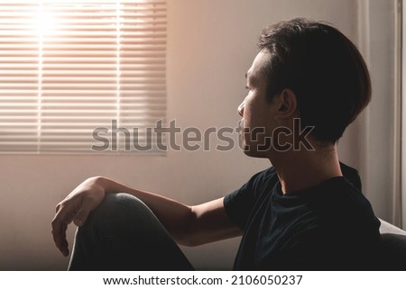 An Asian young man sitting alone near a window on a couch at home office sad and looking down, having problems and hoping for good luck. Loneliness, depression, suffering, financial concept ideas.