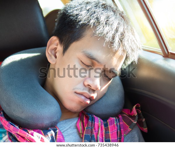 Asian young man sleeping in travel bus. Man\
sleeping in a car travelling on a road trip with blue adult neck\
pillow or neck inflatable pillow. Thai man closed eyes beside car\
windows and sunshine.