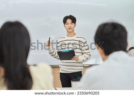 An Asian young man is standing in front of a lecture hall at a university in South Korea, giving a presentation or lecture. In front of him are male and female students.