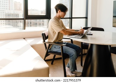 Asian Young Man Studying With Laptop Computer While Sitting By Table In Bright Room At Home