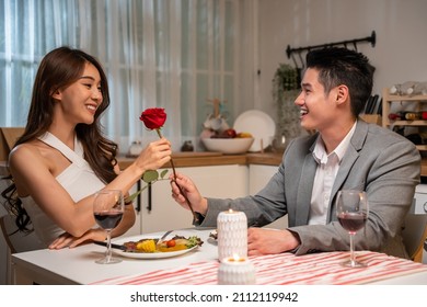 Asian young man surprise and give rose flower to beautiful girlfriend. Attractive romantic new marriage couple man and woman having dinner together to celebrate anniversary and valentines day in house - Shutterstock ID 2112119942