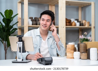 Asian Young Man Talk With Customer On Phone To Check Vase Goods Order. Attractive Business Male Work To Prepare Parcel Boxes, Checking Ecommerce Shipping Online Retail To Sell At Home Warehouse Store.