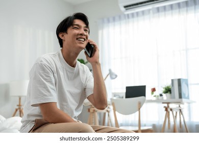 Asian young man talking on mobile phone call on bed at home in morning. Attractive male feeling happy and relax, using smartphone communication with online technology after wake up in bedroom at home. - Powered by Shutterstock