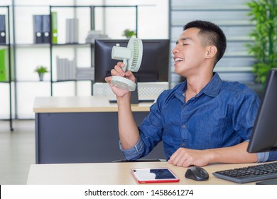 Asian Young Man Using A Small Plastic Fan Blowing To His Face Because Of Hot Weather While Sitting In The Office