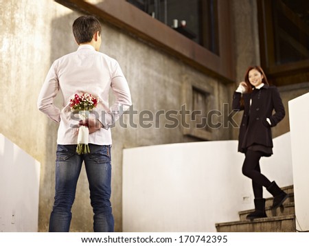 asian young man waiting by the steps with flowers behind back for his girlfriend.
