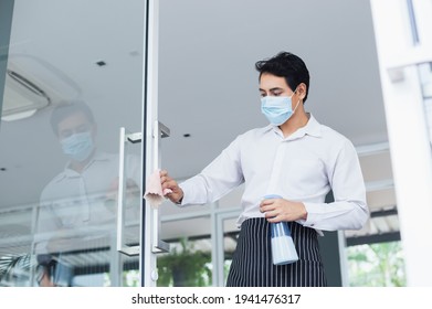 Asian Young Man Wear Face Mask White Shirt Wipe Cleaning Window Glass With Spray Alcohol For Sterilize In Restaurant. Employee Cleaning At Workplace.