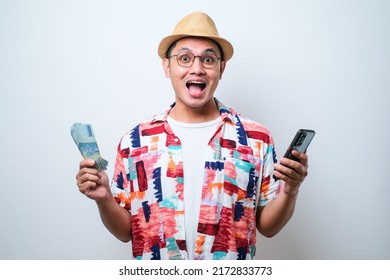 Asian Young Man Wearing Beach Shirt Standing Over White Background Showing Wow Face Expression While Holding Mobile Phone And Paper Money