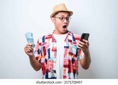 Asian Young Man Wearing Casual Beach Shirt Standing Over White Background Showing Wow Face Expression While Holding Mobile Phone And Paper Money