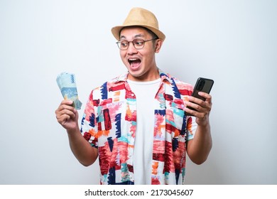 Asian Young Man Wearing Casual Beach Shirt Standing Over White Background Showing Wow Face Expression While Holding Mobile Phone And Paper Money