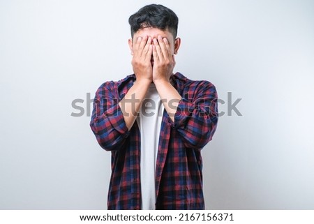 Asian young man wearing casual shirt with sad expression covering face with hands while crying. depression concept over white background