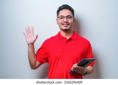 Asian Young Man Wearing Glasses Is Holding A Digital Tablet Standing And Say Hello To Camera Over White Background. Happy Young Man.