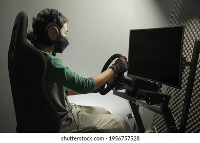 Asian Young Man Wearing Mask In Headphones With Pc Computer Playing Car Racing Video Game At Home And Steering Wheel