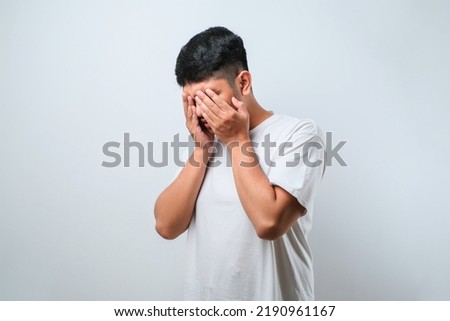 Asian young man wearing white shirt with sad expression covering face with hands while crying. depression concept over white background