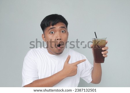 Asian young man wearing a white t-shirt is holding and pointing to iced tea with a wow expression.