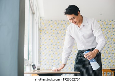 Asian Young Man White Shirt Wipe Cleaning Table With Spray Alcohol For Sterilize In Restaurant. Employee Cleaning At Workplace.