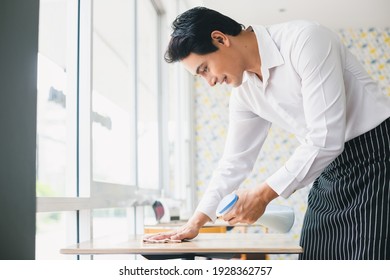 Asian Young Man White Shirt Wipe Cleaning Table With Spray Alcohol For Sterilize In Restaurant. Employee Cleaning At Workplace.