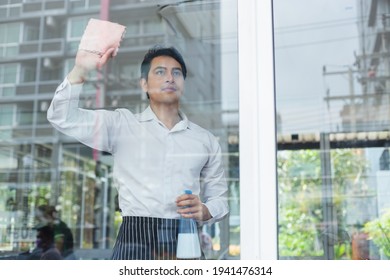 Asian Young Man White Shirt Wipe Cleaning Window Glass With Spray Alcohol For Sterilize In Restaurant. Employee Cleaning At Workplace.